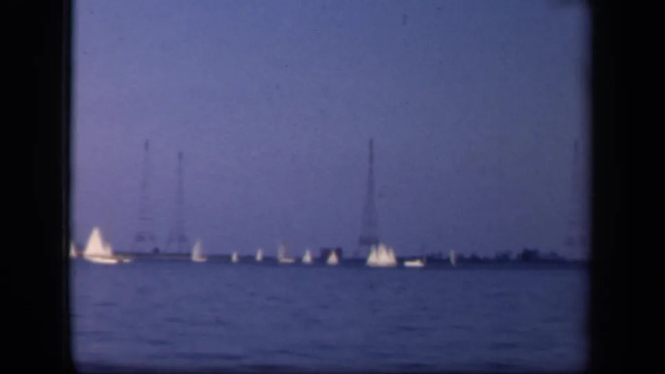Wide home-movie frame of a sailing group on open water in Washington DC, with distant towers faintly visible beyond the boats.
