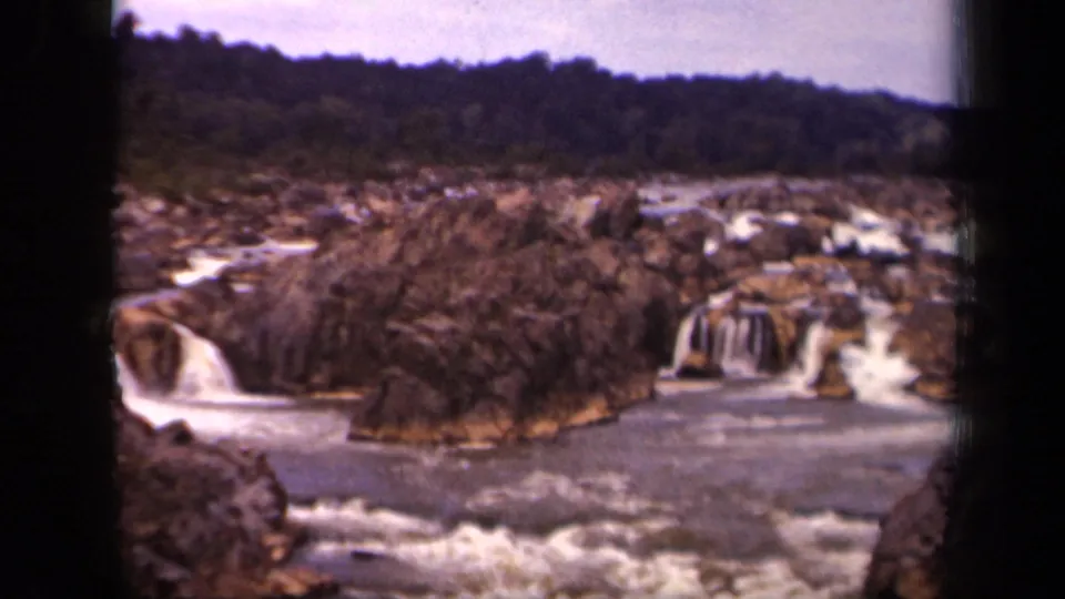 Soft 8mm view from across the river, showing white water, brown rock ledges, brush, and wooded hills in a 1940s Washington-area scene.