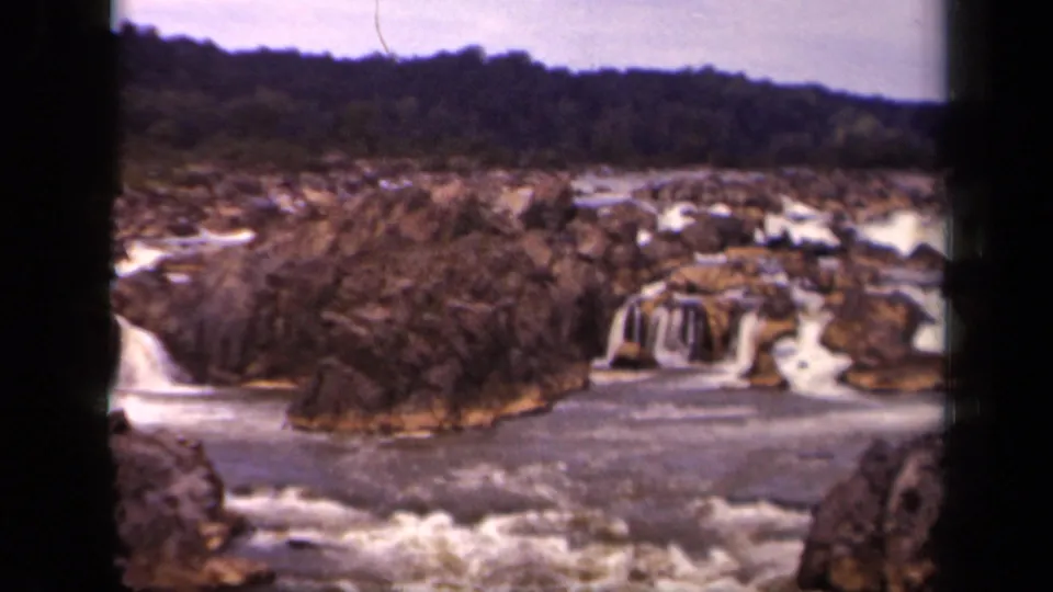 Faded color 8mm frame of a Washington, District of Columbia waterfall landscape, with rocky cascades and a broad river under a muted sky.