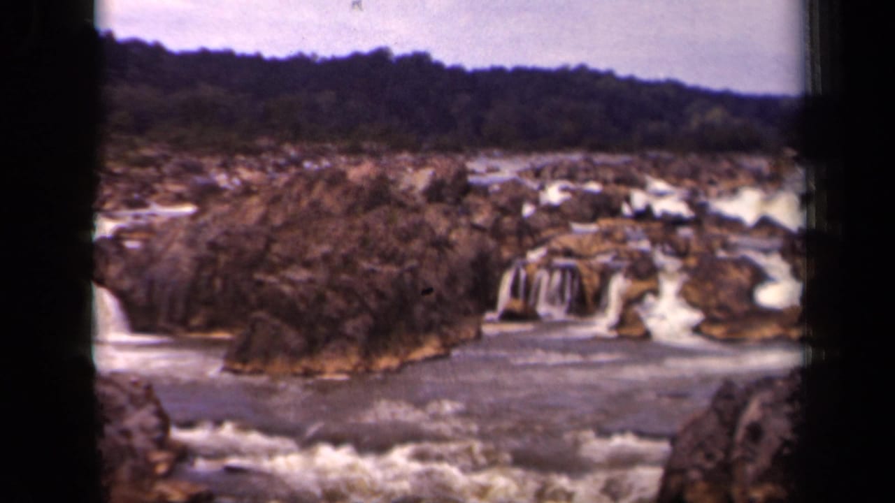 Rocky river waterfall with foamy rapids, wooded ridge, and pale sky.