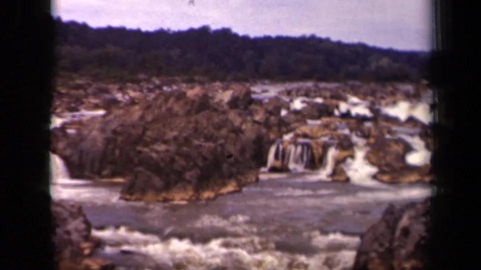 1943 8mm frame from the Washington, DC metadata context showing rocky cascades, foamy river water, trees, and a distant wooded ridge.