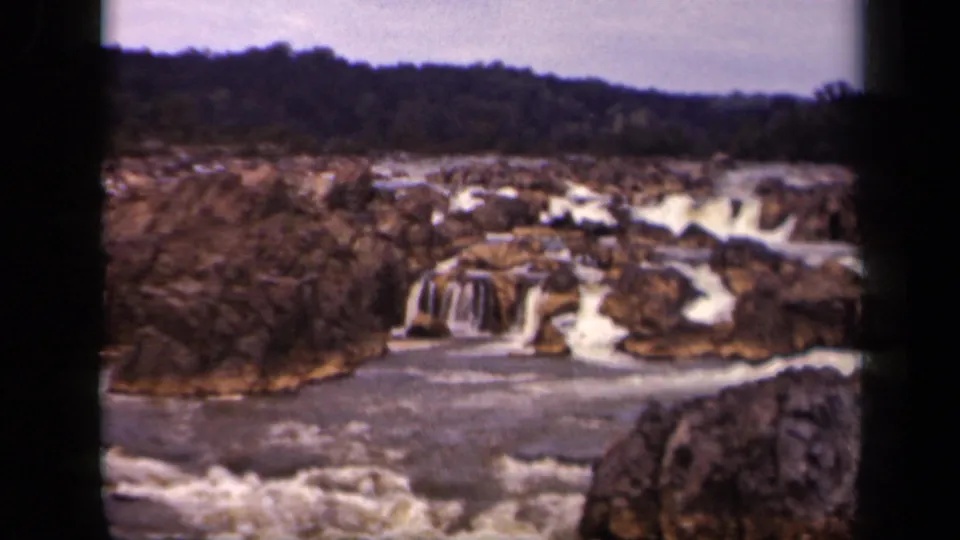 1943 home-movie frame showing foreground rapids, layered rock, low waterfalls, and a wooded ridge under a pale sky.
