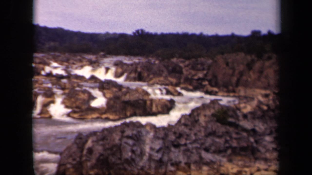 Rushing whitewater flows through dark rocks with a distant tree line under a pale sky.