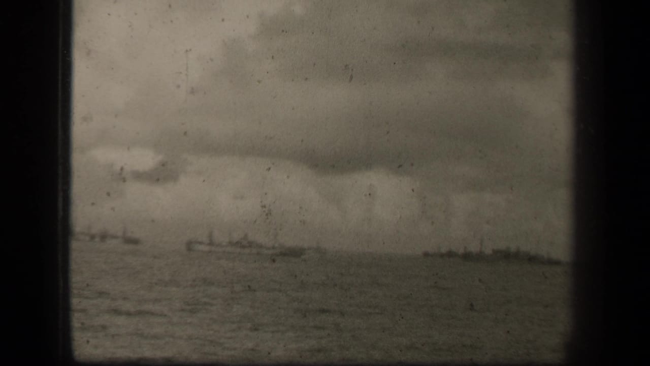 Distant warships on open water beneath a cloudy sky, framed by dark 8mm film edges.