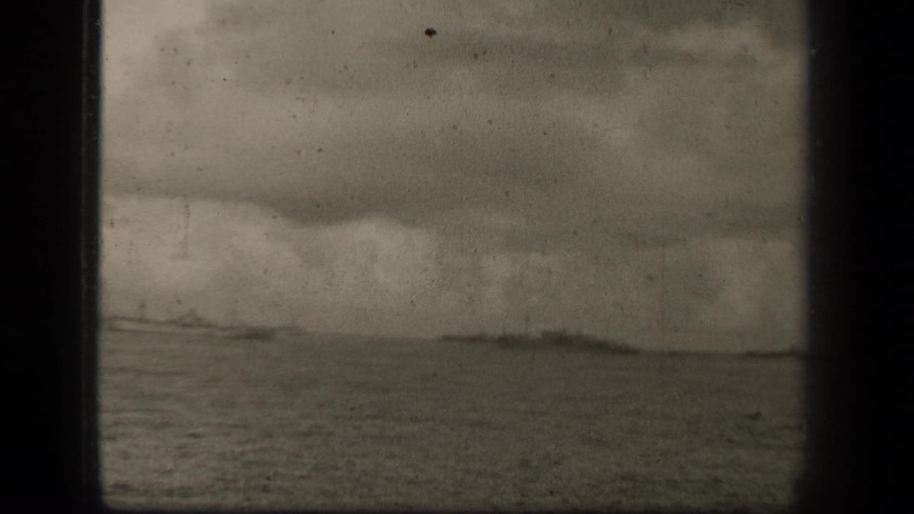Low naval vessels cross a distant horizon on open water under layered clouds.