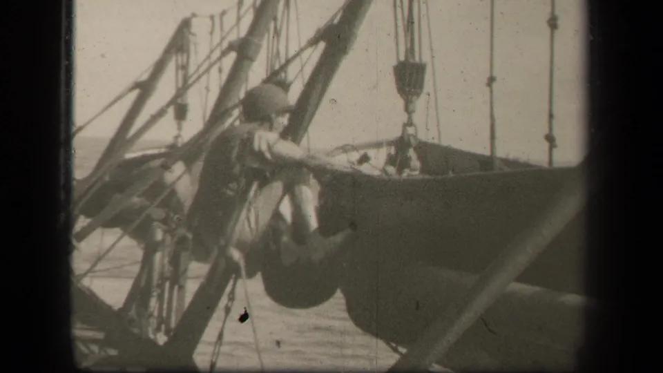 The helmeted serviceman leans forward while climbing over ship structure, framed tightly among rigging and deck hardware in wartime 8mm footage.