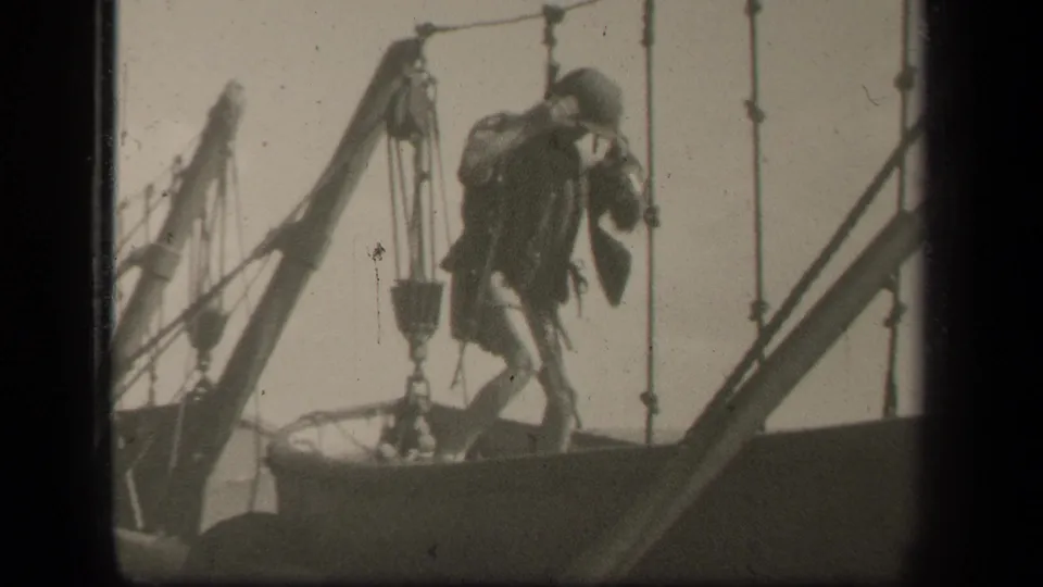 A man in military-style clothing stands on the sailboat deck near ropes and pulley hardware in this 1940s home-movie frame.