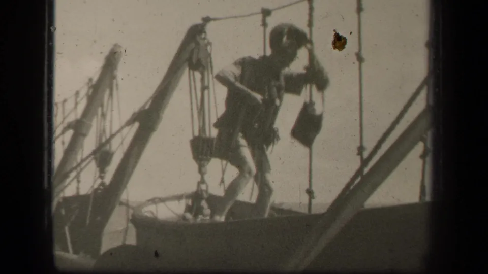 The man stands among mast lines and appears to lift or handle a small item while balanced on the sailboat deck.