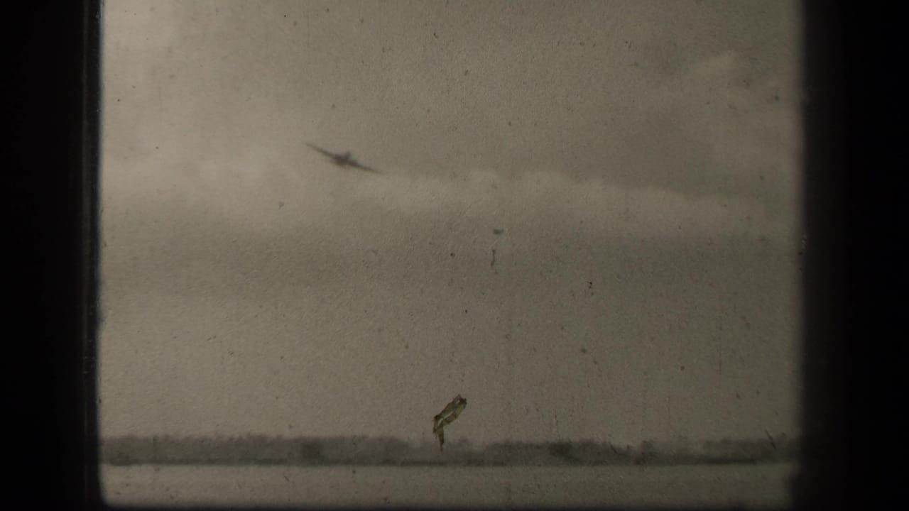 1944 Leyte Gulf 8mm frame showing a small aircraft silhouette above water, with a low shoreline under a cloudy sky.