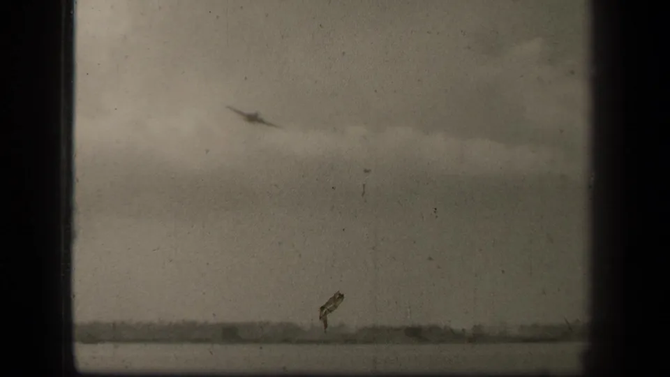1944 Leyte Gulf 8mm frame showing a small aircraft silhouette above water, with a low shoreline under a cloudy sky.