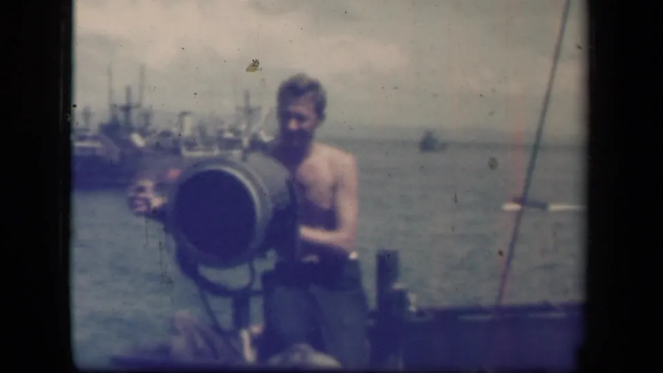 Shirtless man operates or stands by a large signal light on a vessel, with water and ships behind him in the 1944 Leyte Gulf metadata context.