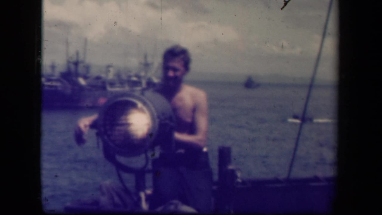 Shirtless sailor stands behind a large searchlight on a vessel with ships in the distance.