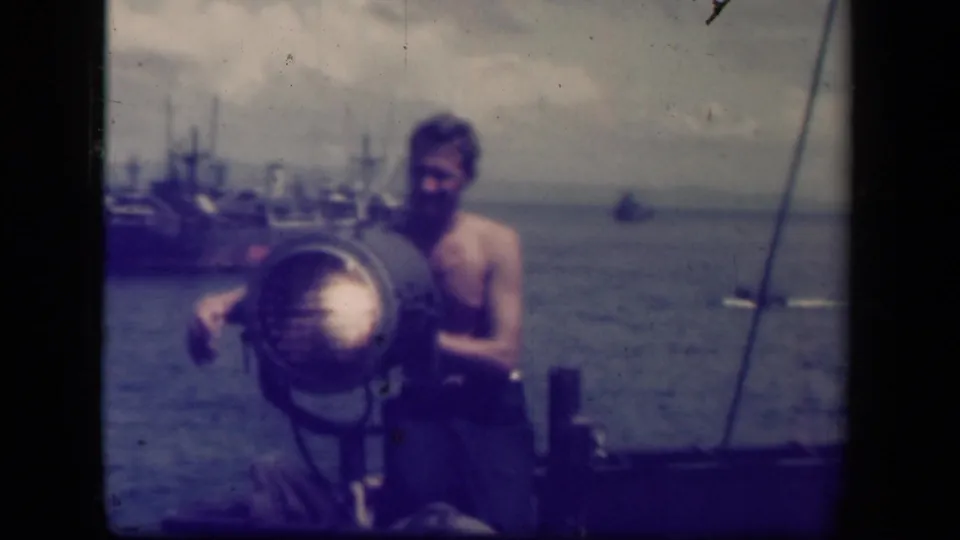 In 1944 Leyte Gulf footage, a shirtless sailor operates a large shipboard searchlight with anchored vessels visible across the water.