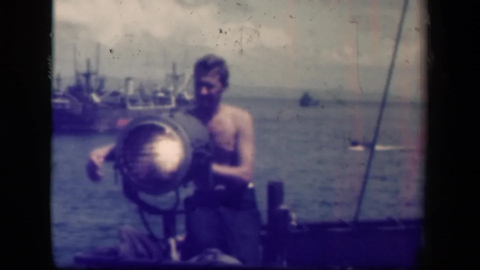 A sailor aboard a naval vessel in the Leyte Gulf footage works at a large searchlight while ships sit in the background.
