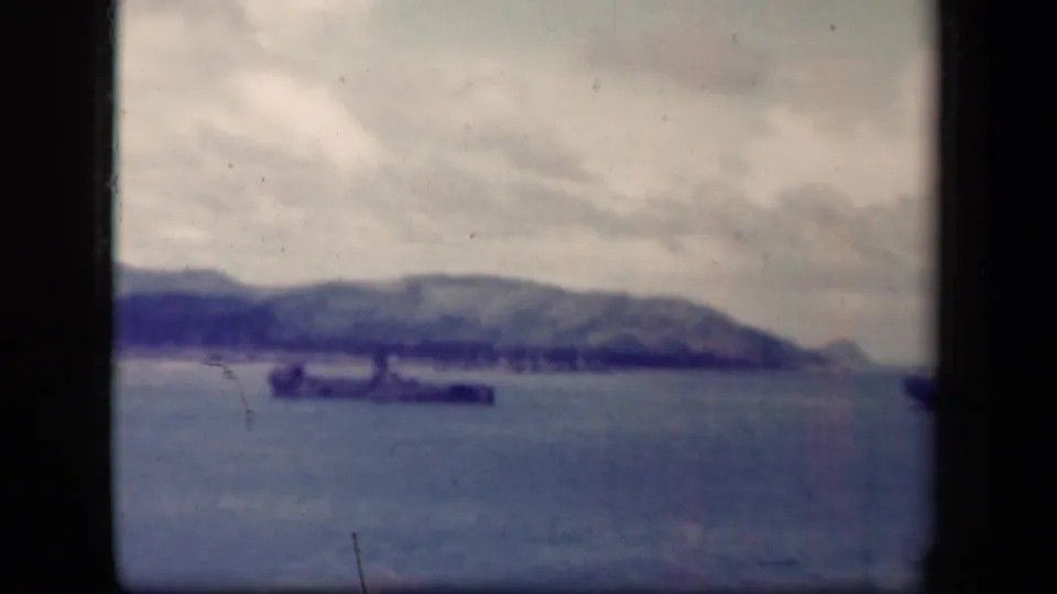 Distant battleship in Leyte Gulf, Philippines, with a mountainous shoreline and cloudy sky, seen through soft 1940s 8mm film texture.