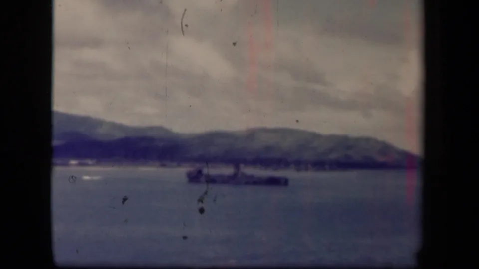 A distant battleship is framed low on the water near a rural peninsula, with muted color, heavy clouds, and visible vertical film wear.