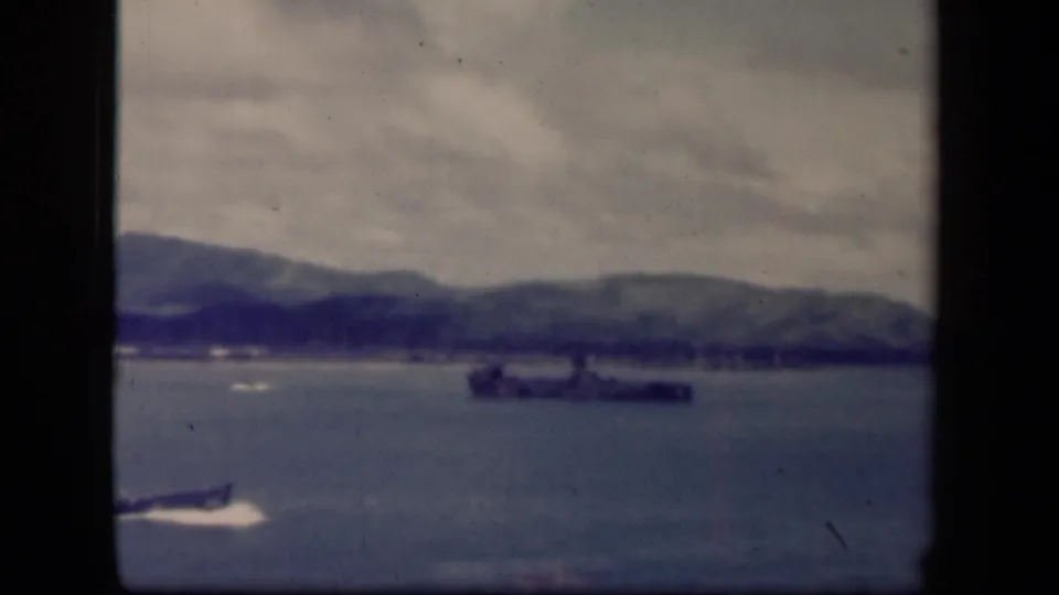 The warship remains offshore in Leyte Gulf, with a small blurred foreground shape, calm water, distant mountains, and worn 8mm film texture.