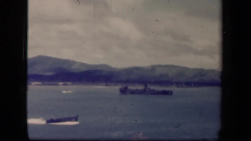 1944 Leyte Gulf 8mm frame showing a small fast boat on blue-gray water, a distant vessel, hazy hills, and dark film borders.