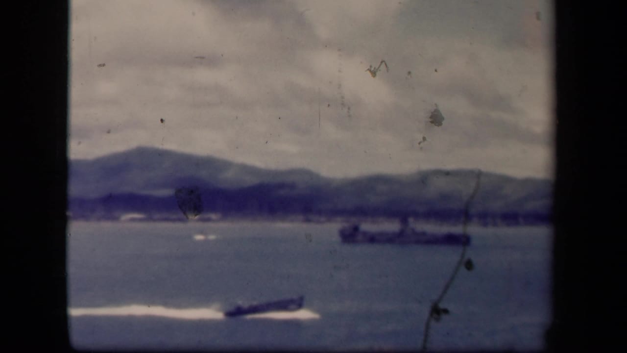 1944 Leyte Gulf 8mm frame with a speedboat and white wake crossing in front of a distant naval vessel and soft shoreline.