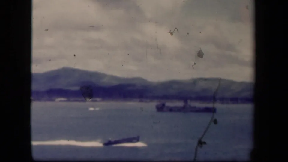 1944 Leyte Gulf 8mm frame with a speedboat and white wake crossing in front of a distant naval vessel and soft shoreline.