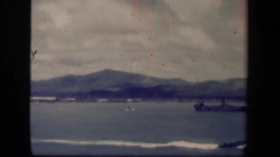 1944 8mm view of Leyte Gulf, Philippines, showing cloudy sky, calm water, distant mountains, shoreline structures, and surf in the foreground.