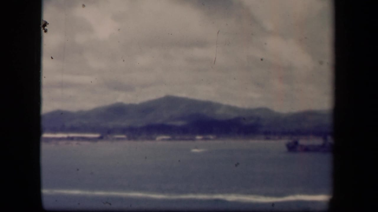 Leyte Gulf shoreline view on 8mm film, with overcast clouds, hazy mountains, small boats, and a larger ship at the right side.