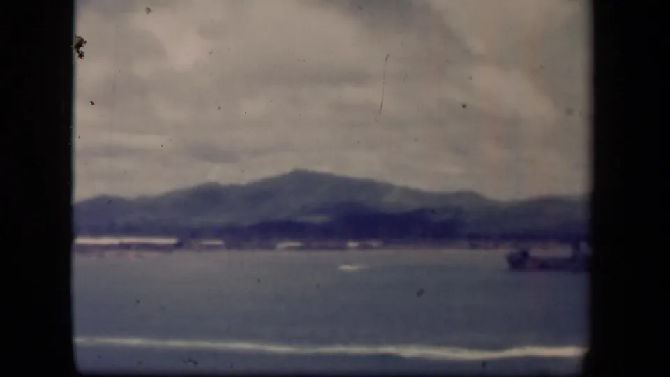 Leyte Gulf shoreline view on 8mm film, with overcast clouds, hazy mountains, small boats, and a larger ship at the right side.