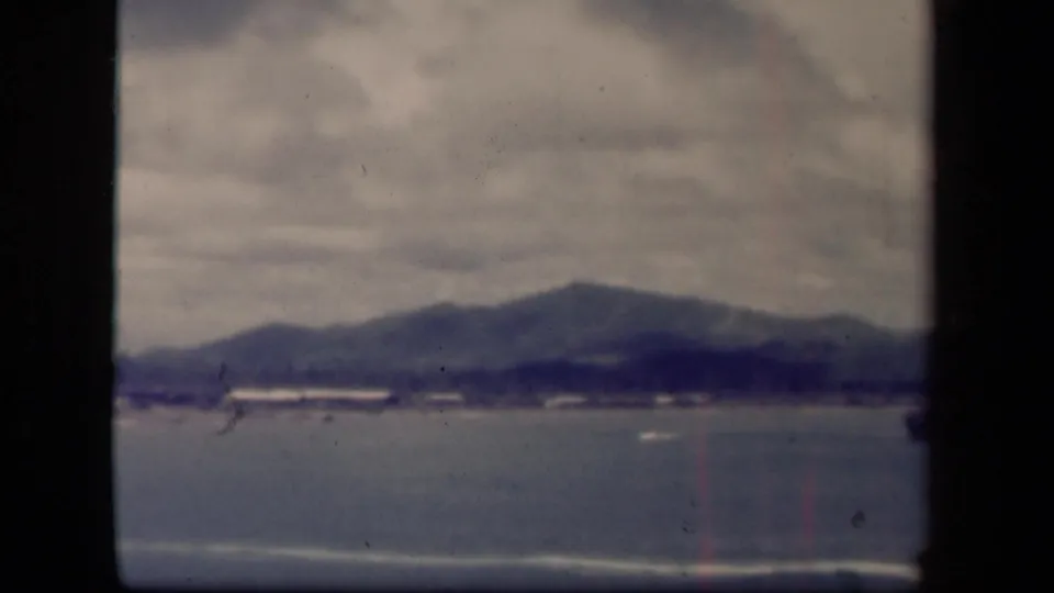 1940s 8mm coastal view of Leyte Gulf with cloudy sky, blue-gray water, distant shoreline buildings, small boats, and soft mountain silhouettes.