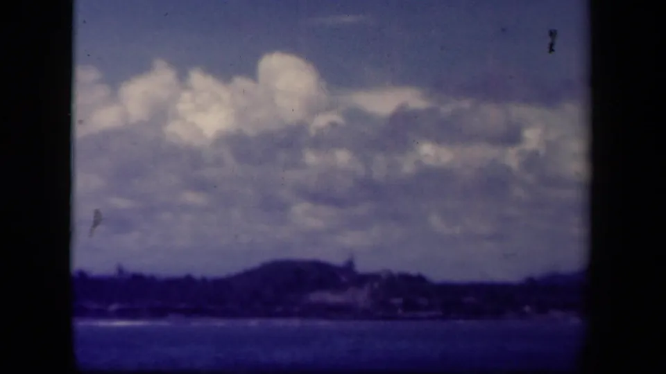 1944 Leyte Gulf 8mm frame showing a distant Philippine shoreline, low hills, and layered clouds above calm water.
