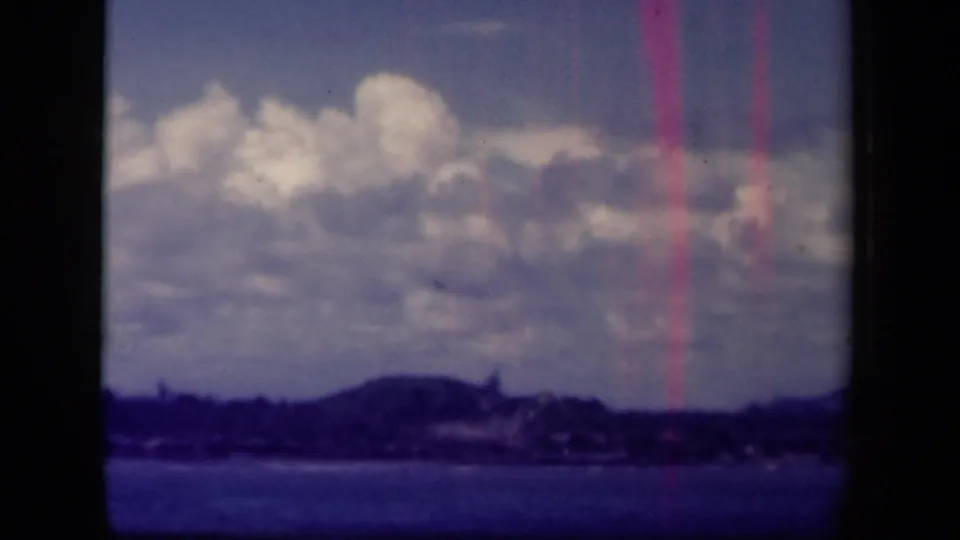 1944 Leyte Gulf 8mm frame showing a hazy shoreline under large clouds across dark blue water.