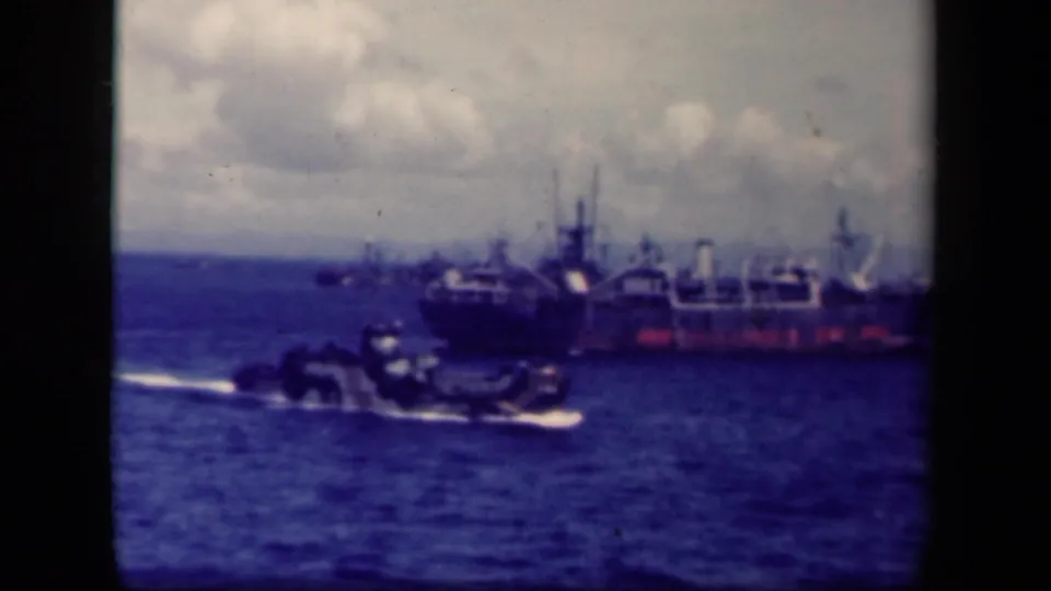 1944 Leyte Gulf 8mm frame showing a large wartime vessel offshore and a smaller craft moving across the foreground water.
