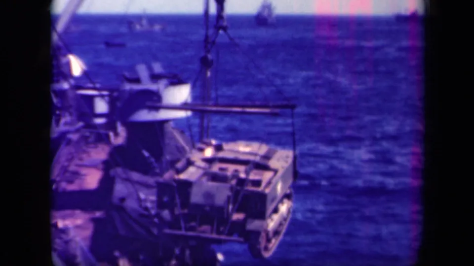Shipboard 1944 Leyte Gulf view of a crane lifting a tracked vehicle above the ocean, with deck railings in the foreground.
