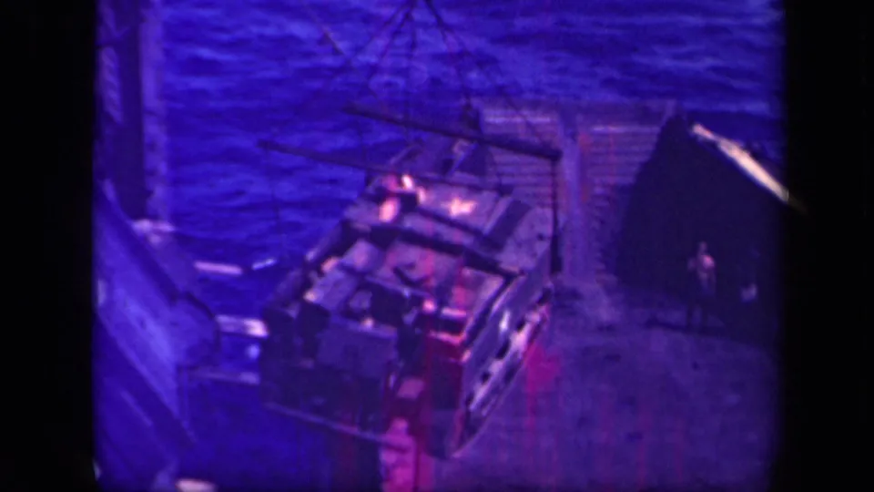 Purple-shifted 8mm frame of a military vehicle being lowered by crane in Leyte Gulf, with figures nearby.