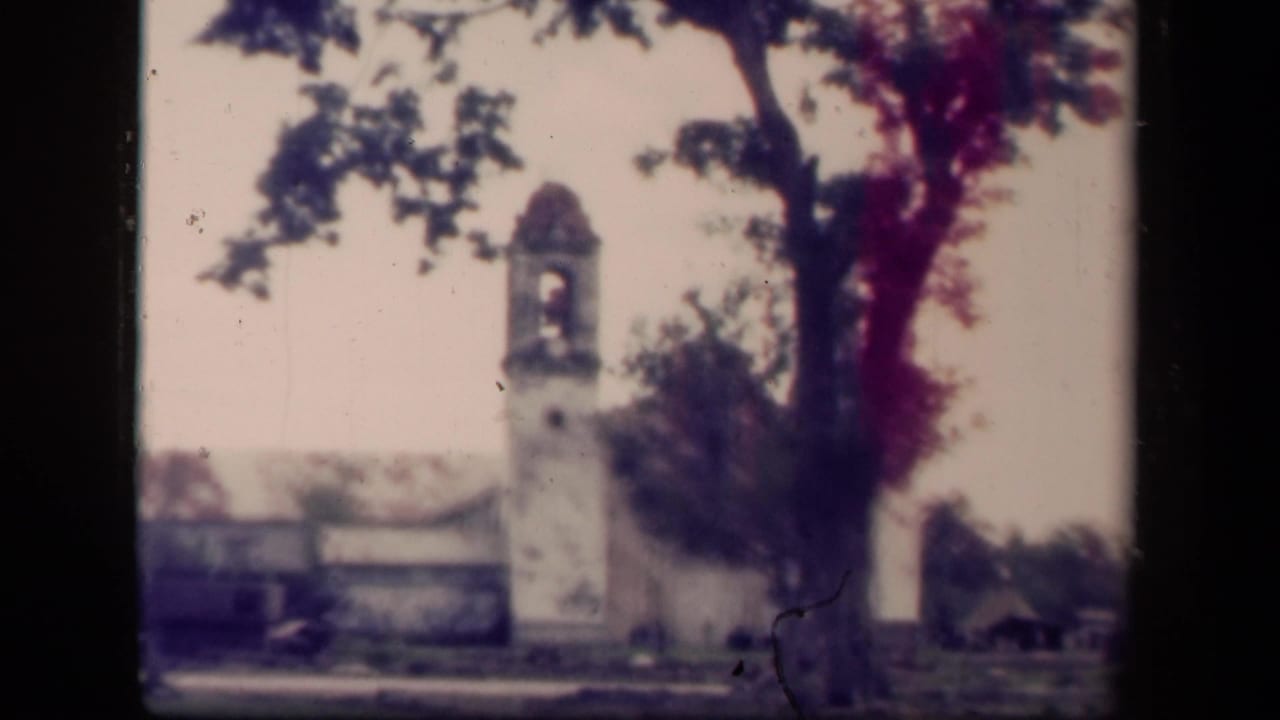 1944 Tacloban home-movie frame showing a white church bell tower with a domed top, framed by a large tree and damaged low buildings.