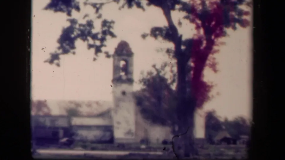 1944 Tacloban home-movie frame showing a white church bell tower with a domed top, framed by a large tree and damaged low buildings.