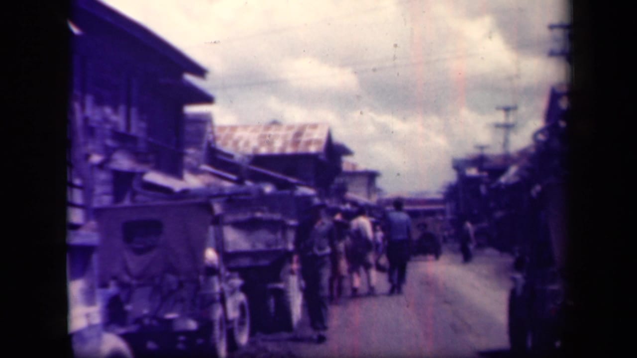 Wartime-era Leyte Gulf street view with men walking beside older vehicles, carts, low buildings, and utility poles under a cloudy sky.