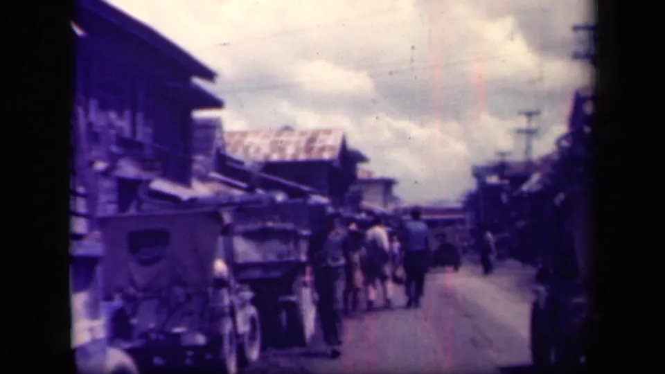 Wartime-era Leyte Gulf street view with men walking beside older vehicles, carts, low buildings, and utility poles under a cloudy sky.