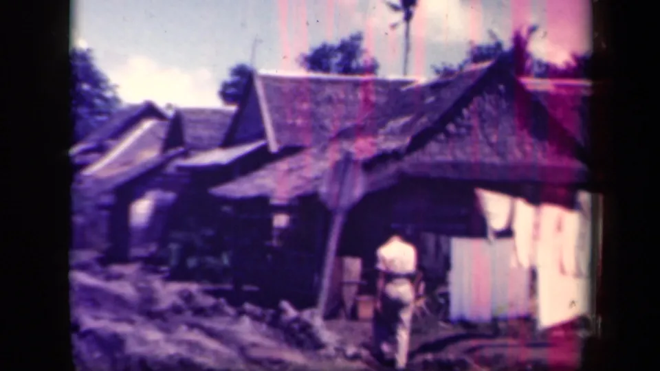 A person stands near a thatched-roof dwelling at the roadside, with village houses and a muddy track visible in the 1940s Leyte Gulf footage.