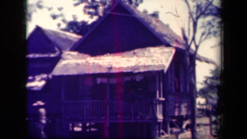 1944 Leyte Gulf home-movie frame showing raised wooden dwellings with thatched roofs, porch railings, trees, and a distant person.