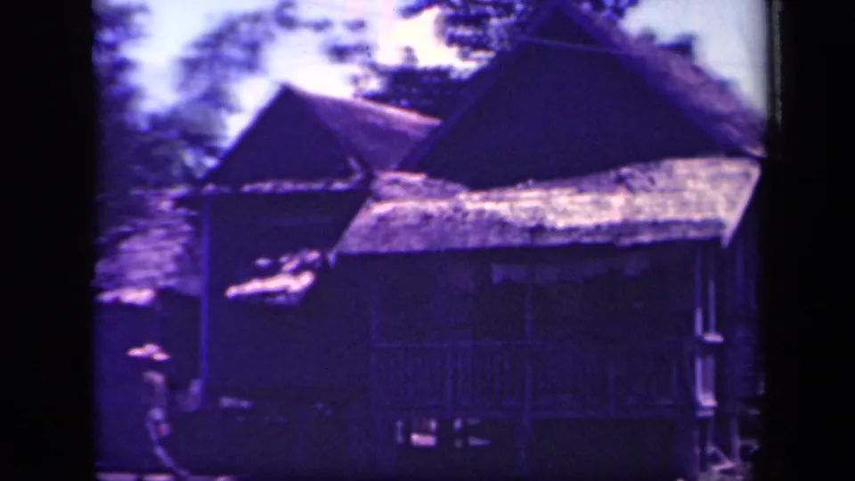 1944 Leyte Gulf frame with darkened stilt houses, thatched roofs, trees, and a distant person near the foreground.