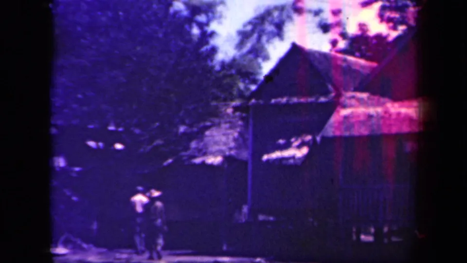 1944 Leyte Gulf frame showing a thatched stilt house, dense trees, and two distant people walking near the lower left.