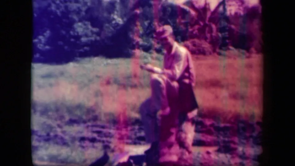 A seated man in a tropical Leyte Gulf field appears to read or write, with palms and brush behind him in faded 8mm color.