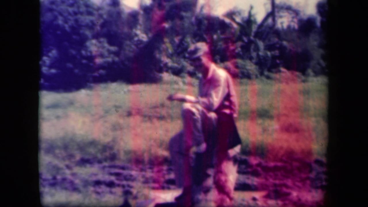 Uniformed man sits on a log in a grassy tropical field with foliage behind him.