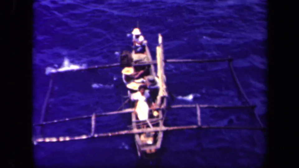 The high-angle 8mm frame shows men in hats paddling a traditional outrigger canoe over dark blue water in the Philippines.