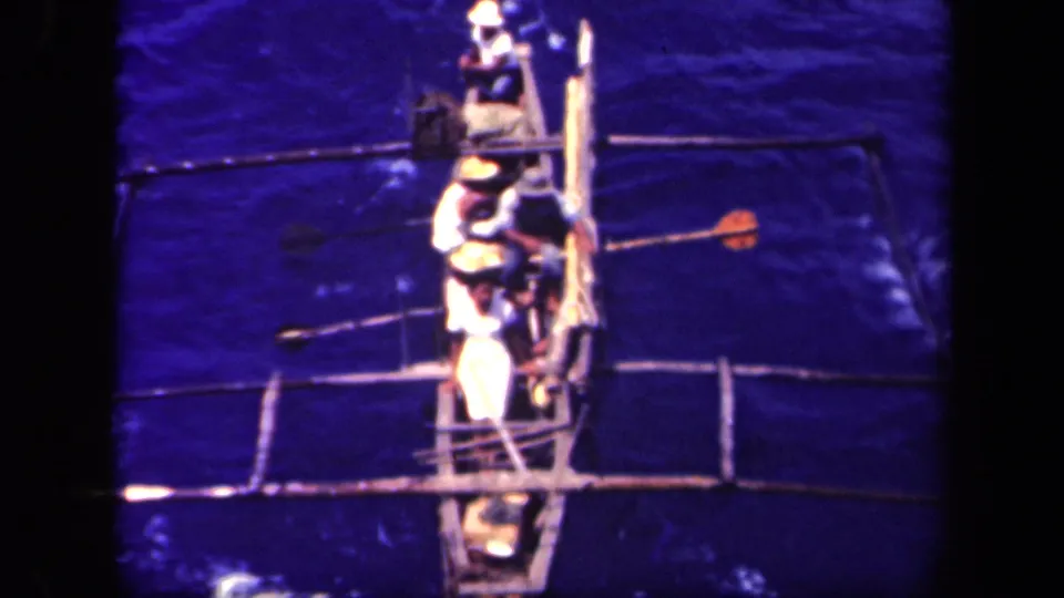 High-angle 8mm frame of men in a narrow outrigger canoe, with paddles and crossbeams visible over blue water.