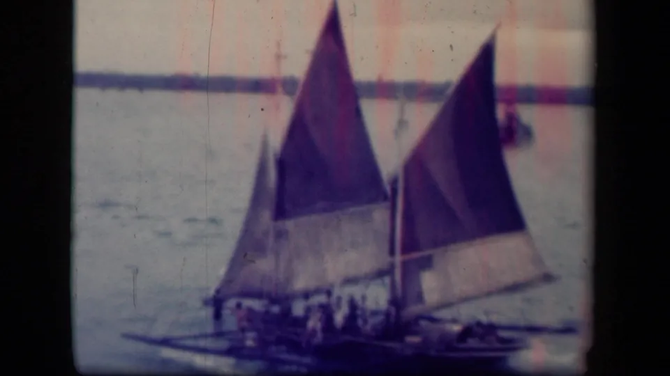 The sailboat continues across Leyte Gulf, with people aboard beneath two raised triangular sails and a low shoreline beyond.