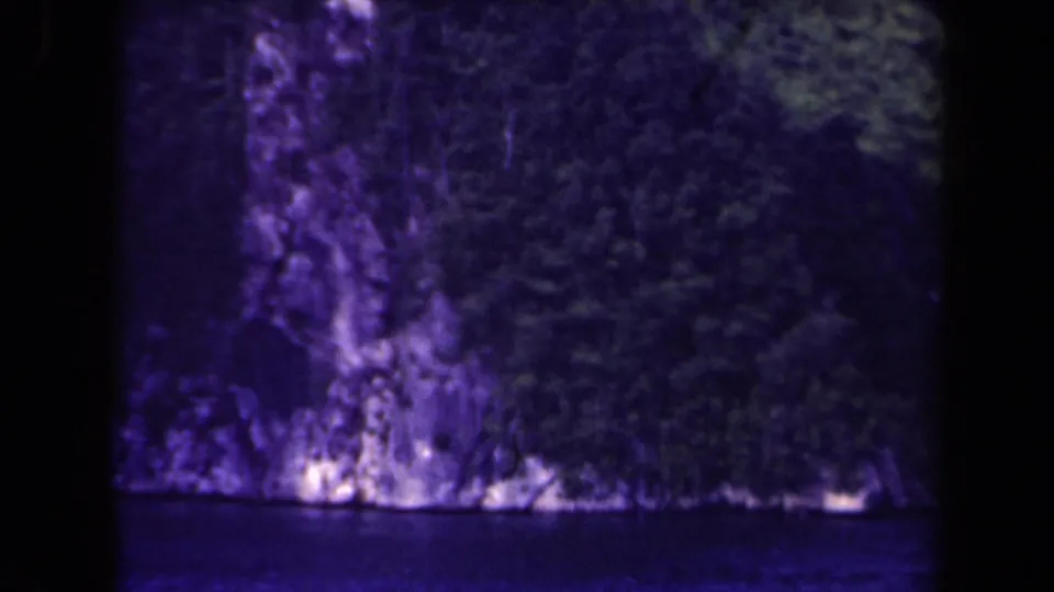 Papua New Guinea coastal cliff covered in dense vegetation, seen across dark water on aged 8mm film.