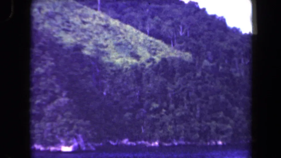 A sloping Papua New Guinea forested hillside appears in a purple-tinted 1944 8mm frame, with water visible at the bottom.