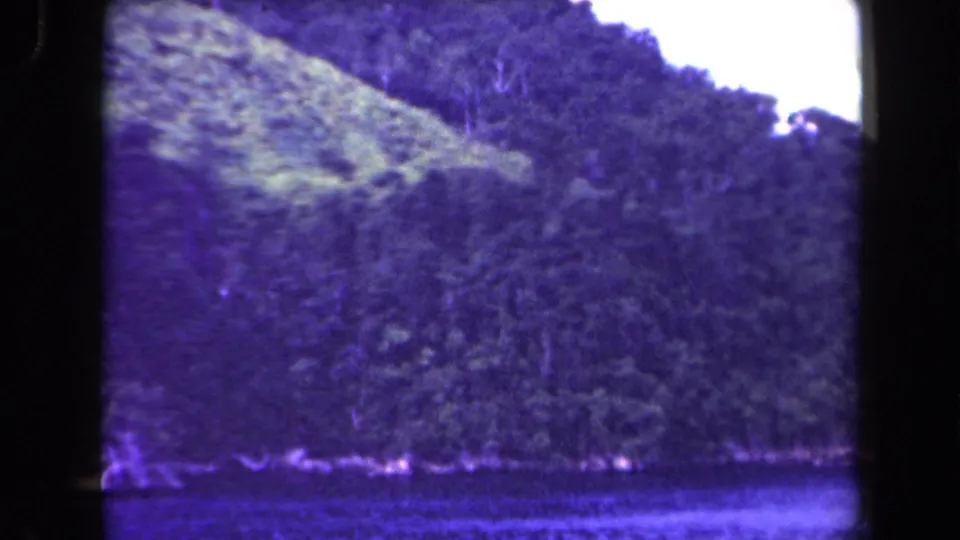 Final 1944 8mm frame of a forested Papua New Guinea mountainside across water, with soft grain, purple cast, and heavy frame-edge shadows.