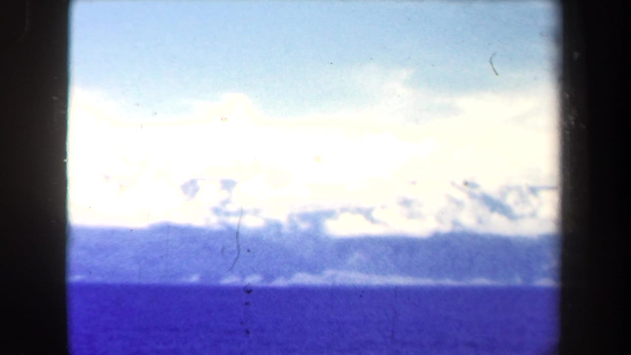 1944 Papua New Guinea 8mm aerial view of blue water, distant mountain ridges, and cloud cover within a dark film frame.